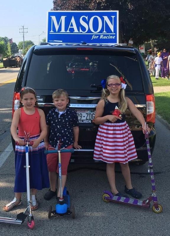Three Mason children with scooters by a "Mason for Racine" van at the Fourth of July parade